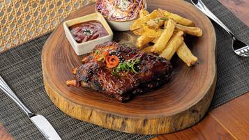 A wooden serving board with a grilled meat dish, French fries, and a side salad on a table with a woven chair and a metal fork.