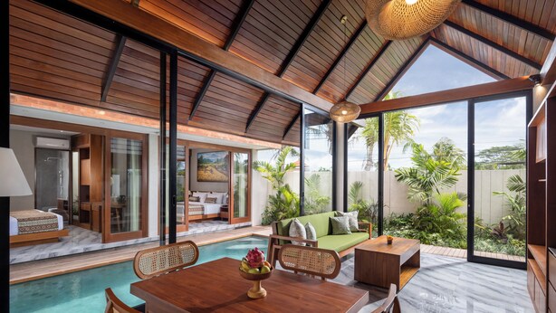 A modern, open-plan living area with a pool, wooden furniture, and large windows offering a view of the outdoors. - Unagi Mas Villas by Emana (Mas)