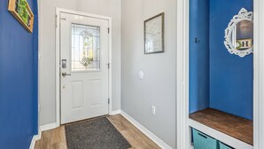 A hallway with a white door, a blue wall, a wooden shelf, and a mirror with a letter 'H'.
