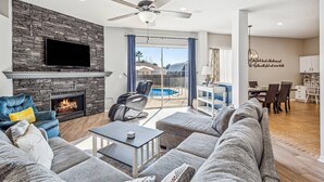 A spacious living room with a stone fireplace, a flat-screen TV, a ceiling fan, and a pool view.