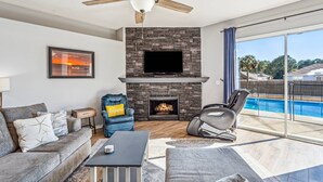 A modern living room with a stone fireplace, a ceiling fan, a large TV, and a pool view.