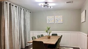Ruokailutila, jossa on puinen pöytä, vihreät tuolit, ruukkukasvi ja kuviolliset verhot.