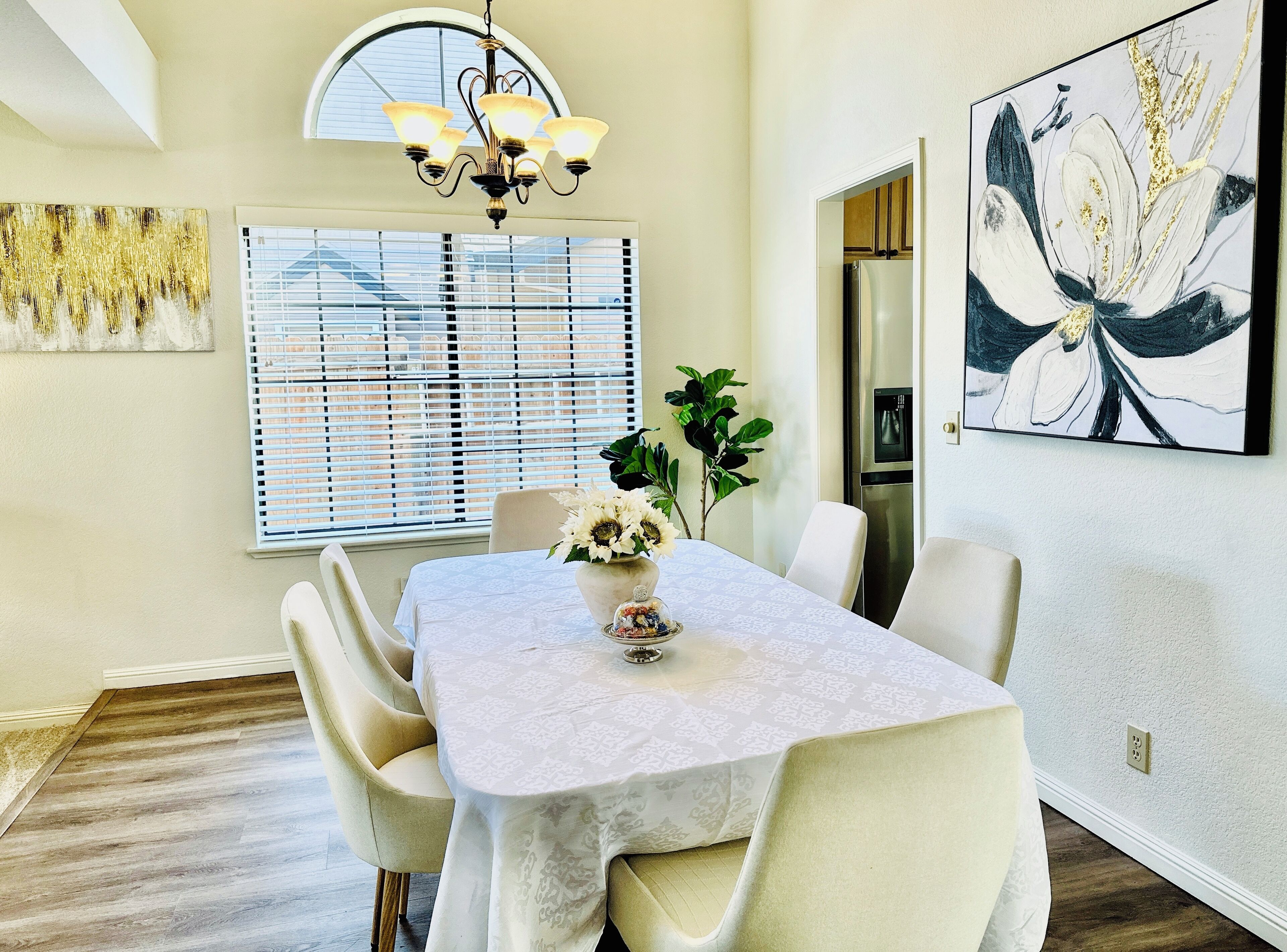 A dining area with a table set for four, a chandelier, a window with blinds, and a large abstract painting on the wall.