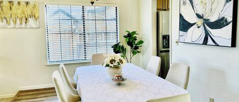 A dining area with a table set for four, a chandelier, a window with blinds, and a large abstract painting on the wall.
