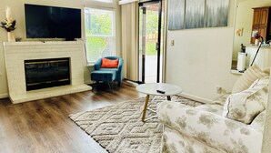 Una sala de estar con chimenea, televisor, un sillón azul, un sofá estampado y vistas al exterior.
