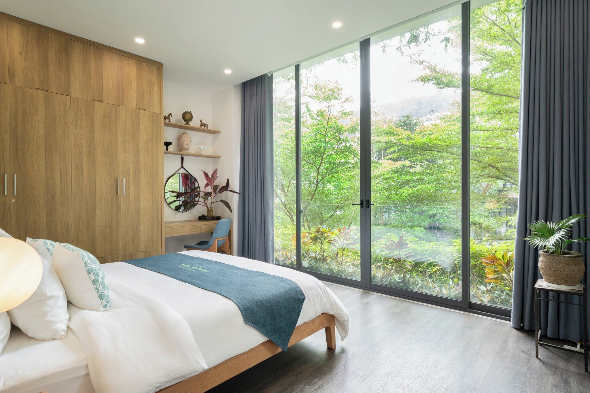 Une chambre moderne avec un grand lit, une armoire en bois et une vue sur une végétation luxuriante grâce à de grandes fenêtres du sol au plafond.