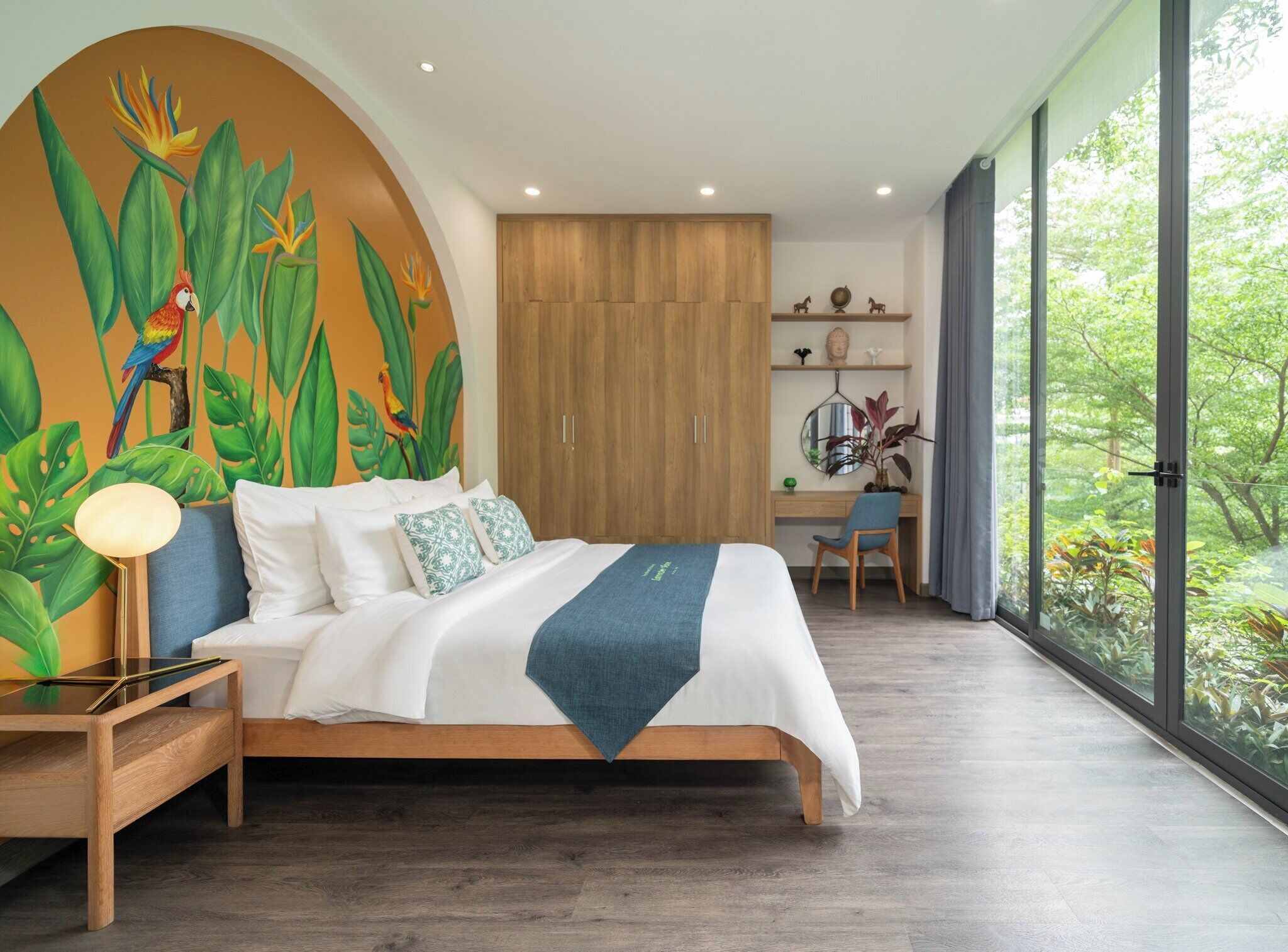 A modern bedroom with a large bed, a desk, and a view of greenery through large windows.