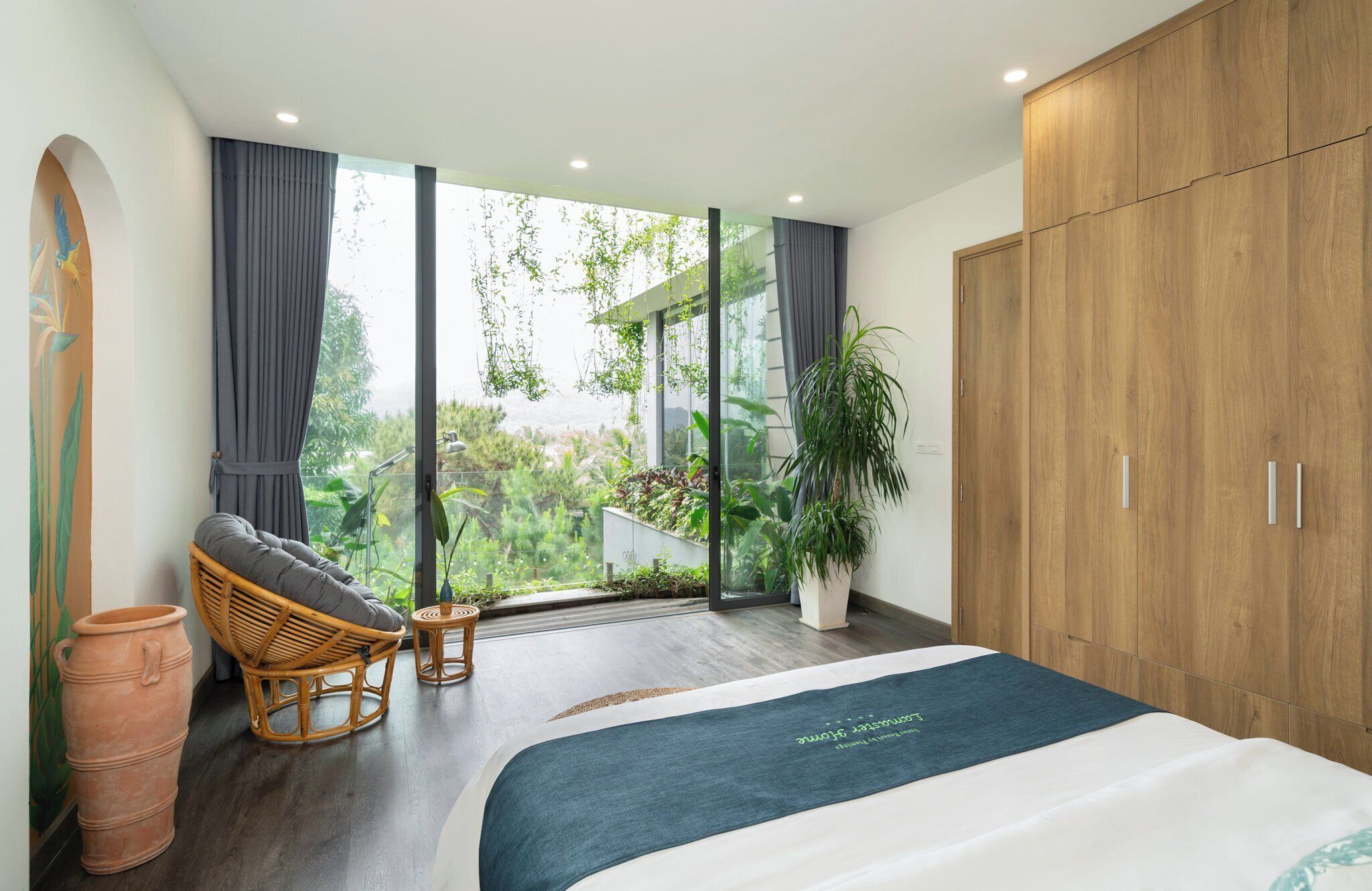 Une chambre avec un grand lit, une armoire en bois, un fauteuil en osier, une petite table et une vue sur la verdure grâce à de grandes fenêtres.