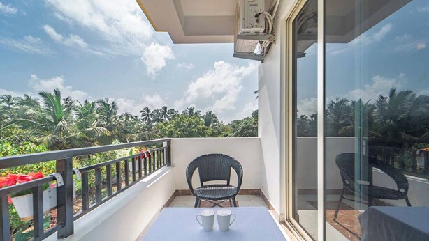 A balcony with a table, two chairs, and a view of palm trees and greenery. - Artham Guesthouse (Morjim)