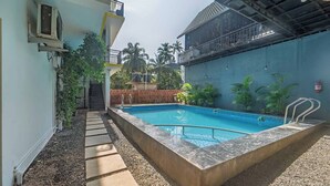 A swimming pool with a tiled deck, surrounded by a low brick wall and greenery. - Artham Guesthouse (Morjim)