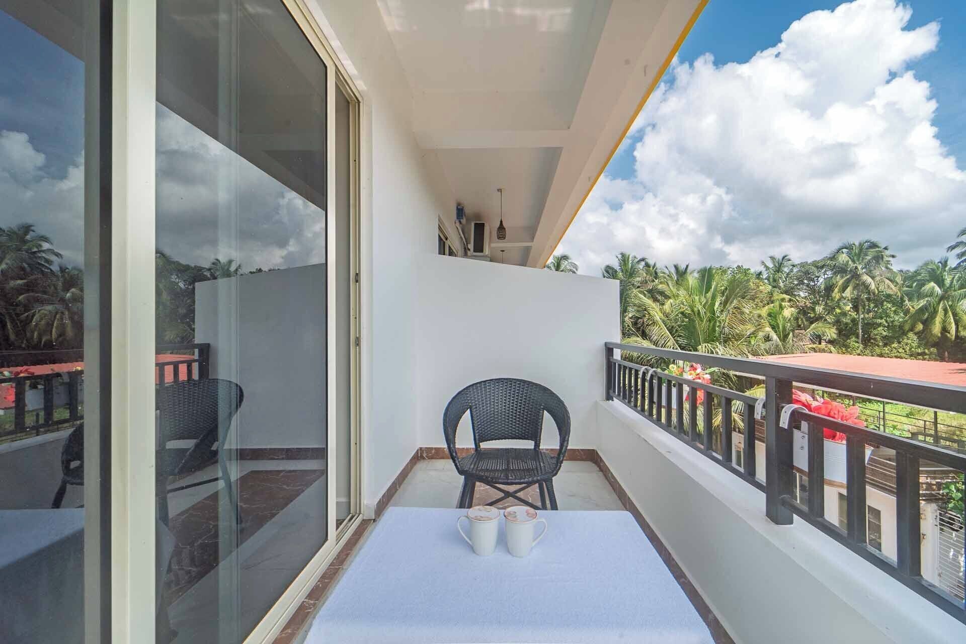 Ein Balkon mit einem weißen Tisch, zwei weißen Tassen, einem schwarzen Korbsessel und Blick auf Palmen sowie einen bewölkten Himmel.