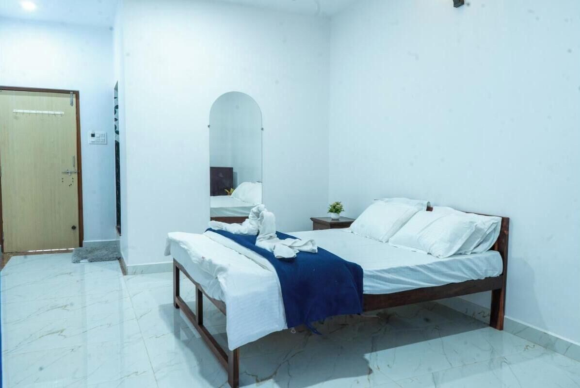 A bedroom with a wooden bed, white bedding, a blue throw, a small bedside table with a plant, and a mirror on the wall.