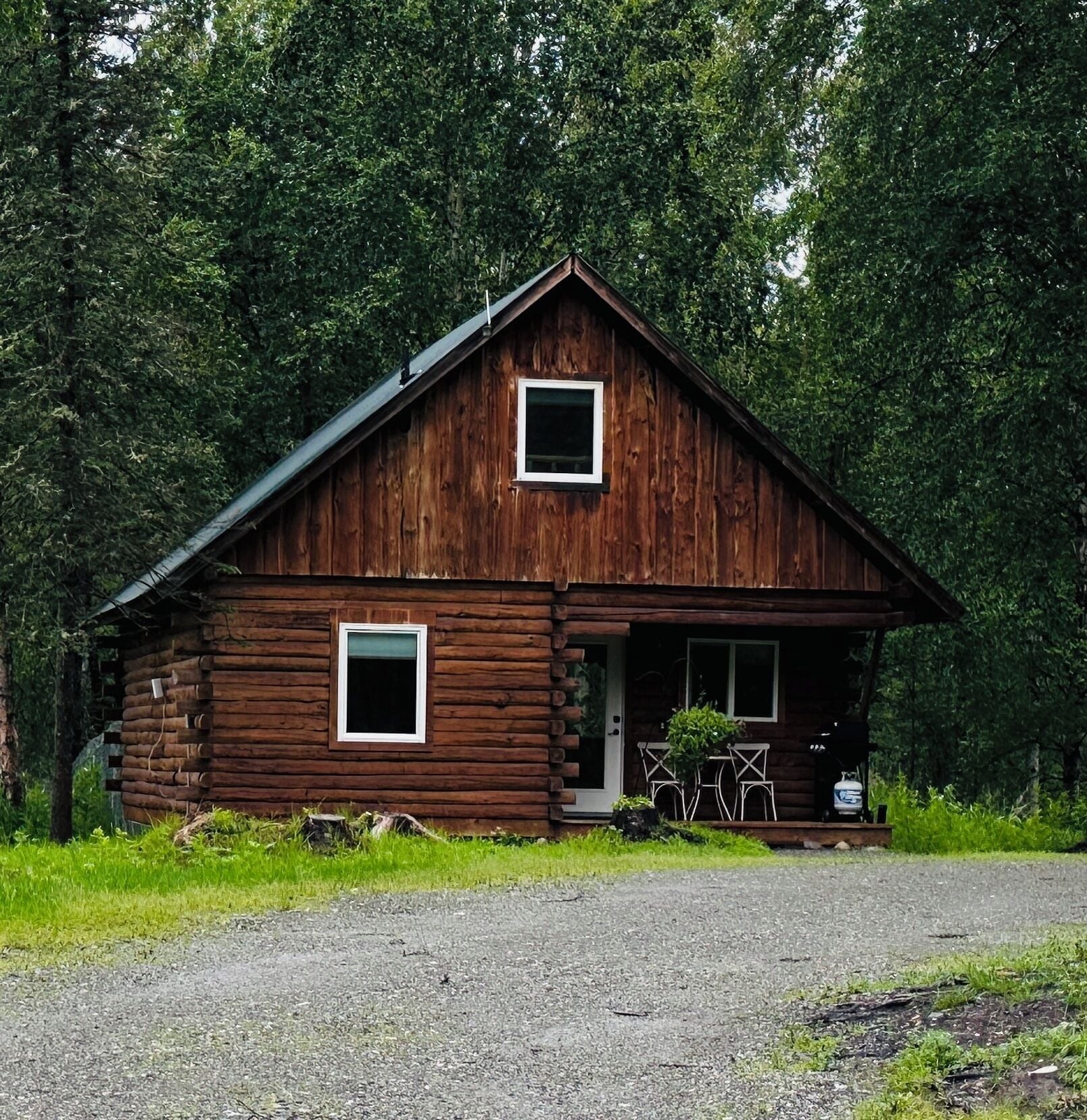 A wooden cabin with a porch and two windows, surrounded by dense forest.