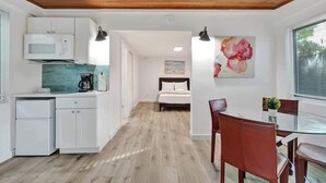 A modern kitchen with white cabinets, a microwave, and a coffee maker. There is a dining area with a glass table and red chairs. A bed is visible in the background. - Tropical Studio + Pool | Riverside Ft Lauderdale (Fort Lauderdale)