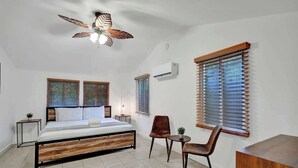 Une chambre à coucher comprenant un lit, un ventilateur de plafond, deux fenêtres avec stores, une table de chevet, une chaise et une petite table.
