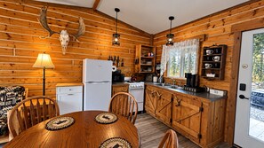 A cabin interior with a wooden dining table, chairs, a refrigerator, a stove, and a kitchen area with wooden cabinets and a sink. - Cozy, pet-friendly, 2-bedroom cabin in beautiful Palmer, Alaska (Palmer)