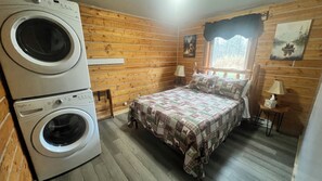 A room with a bed, a washing machine, wooden walls, a window with a view, and a painting on the wall. - Cozy, pet-friendly, 2-bedroom cabin in beautiful Palmer, Alaska (Palmer)