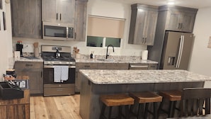 Une cuisine moderne avec des meubles en bois, un plan de travail en granit, un four encastré, un réfrigérateur et un coin bar avec des tabourets.