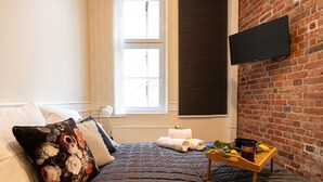 A bedroom with a bed, a window, a TV mounted on a brick wall, and a tray with items on the floor. - Arcano | Studio dla Dwojga | Przytulne wnętrze (Bydgoszcz)