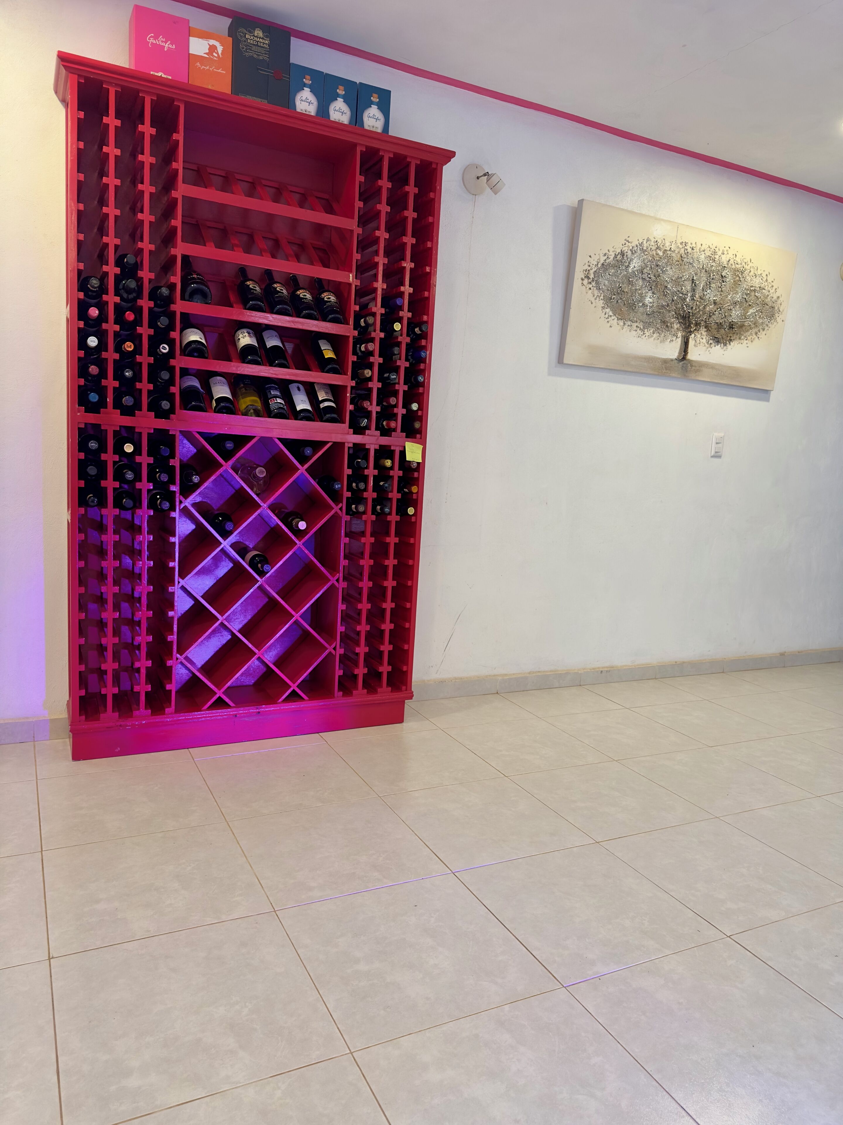 Uma estante para vinho tinto com várias prateleiras, um quadro de uma árvore na parede e um chão em azulejos.