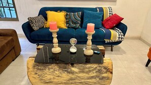 Una sala de estar con un sofá azul, una mesa de centro de madera con dos velas y una hora de arena, y un sillón marrón.