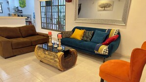 Una sala de estar con un sofá marrón, un sofá verde azulado con cojines coloridos, una mesa de centro de madera con velas y un sillón naranja.