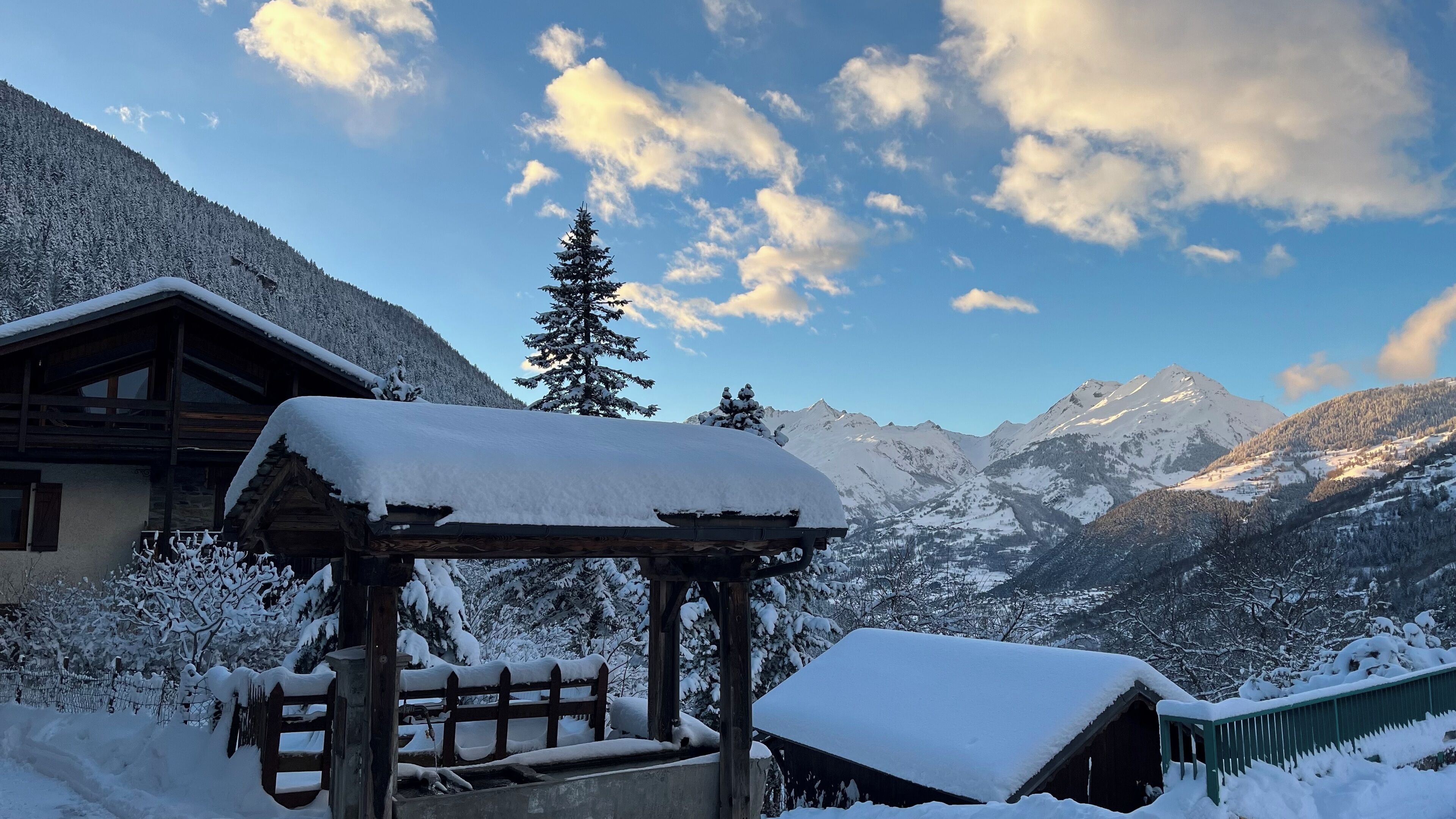 Una cabaña de madera con techo nevado, cerca una valla de madera, y vista a montañas cubiertas de nieve bajo un cielo parcialmente nublado.