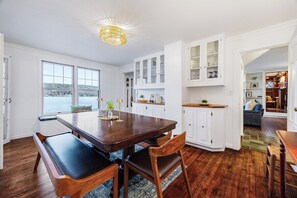 Un espace repas comprenant une table et des chaises en bois, des meubles blancs aux portes vitrées et une vue sur l’extérieur.