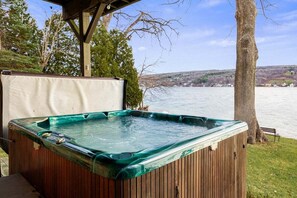 Un bain à remous avec une structure extérieure en bois et un couvercle en verre, installé à l’extérieur et offrant une vue sur un lac et des collines en arrière-plan.