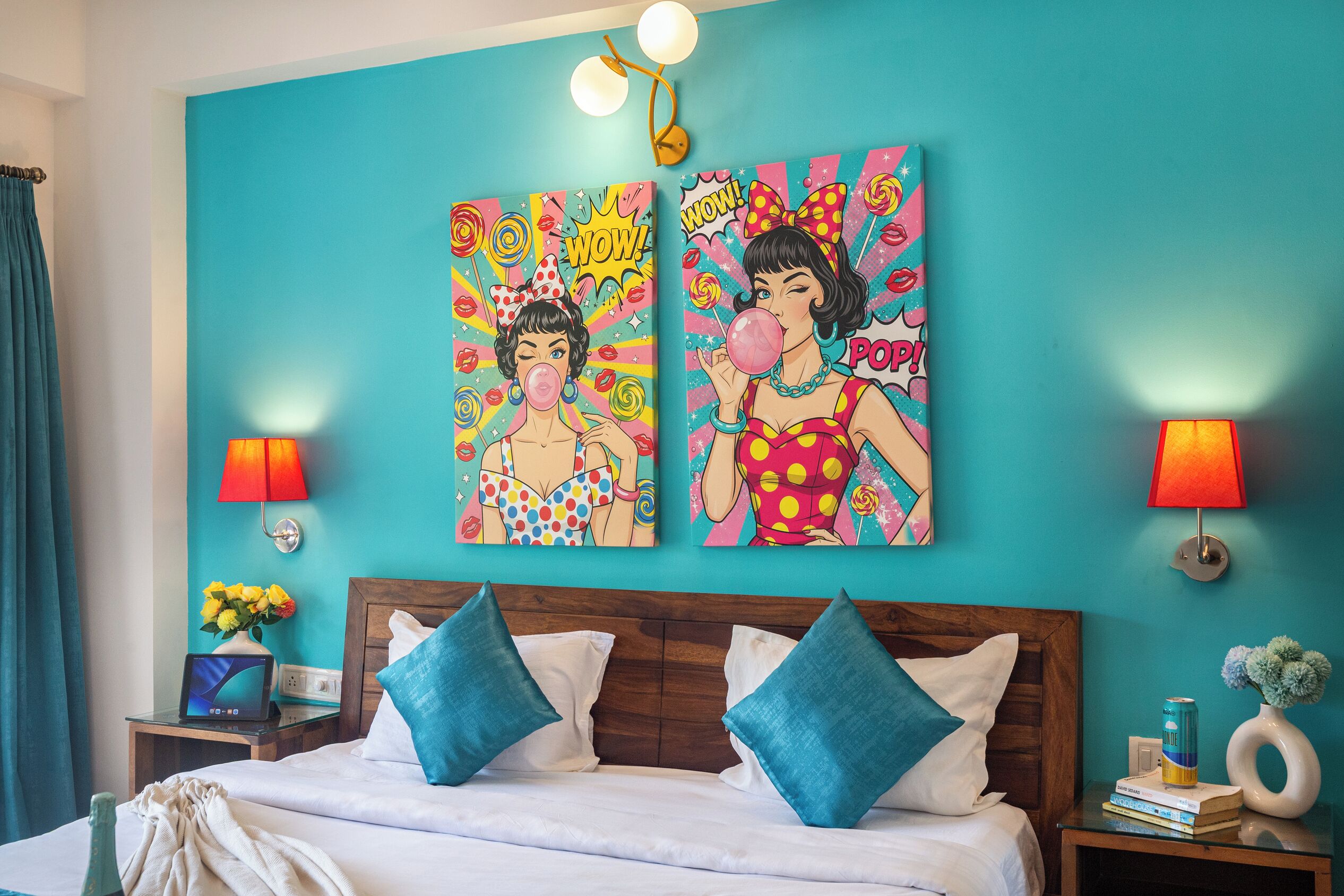 A hotel room with a teal wall, a wooden headboard, two blue pillows, a laptop on a bedside table, and two wall-mounted lamps.