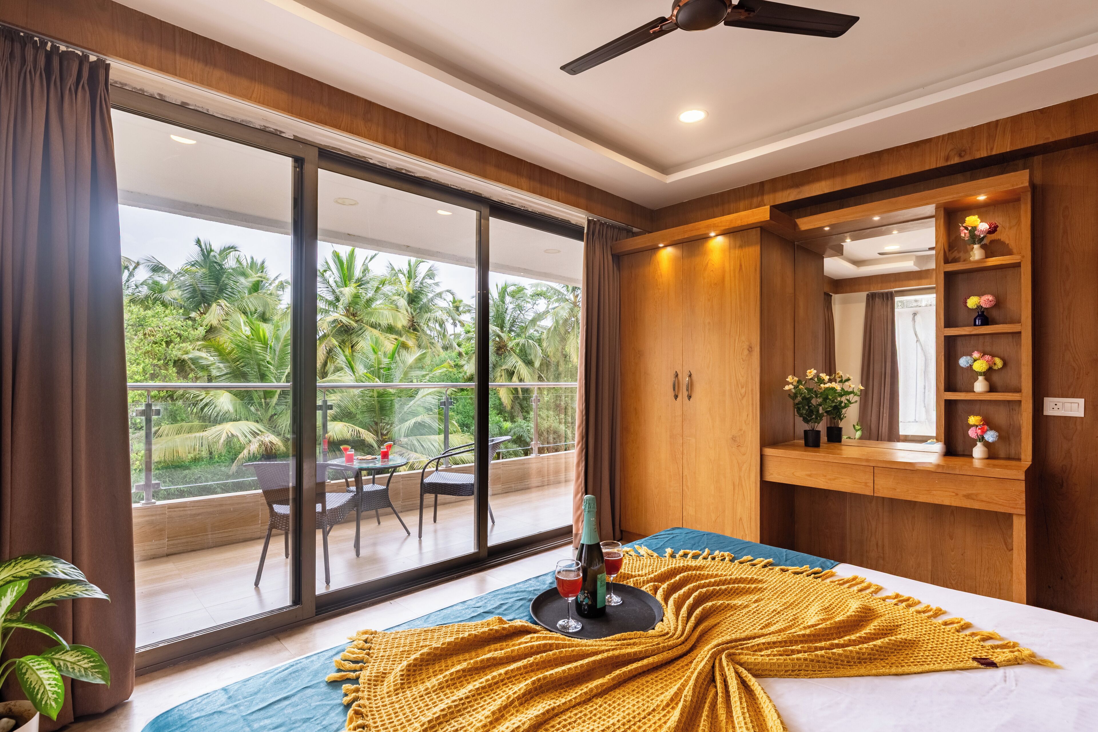 A bedroom with a large sliding glass door to a balcony, a bed with a yellow blanket, a bedside table with a bottle and glasses, and a wooden vanity with a mirror and shelves.
