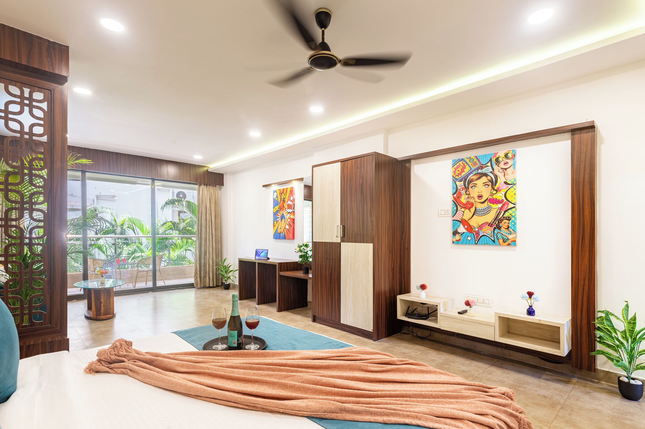 A modern hotel room with a bed, a ceiling fan, a desk, a window with a view of greenery, and a painting on the wall.