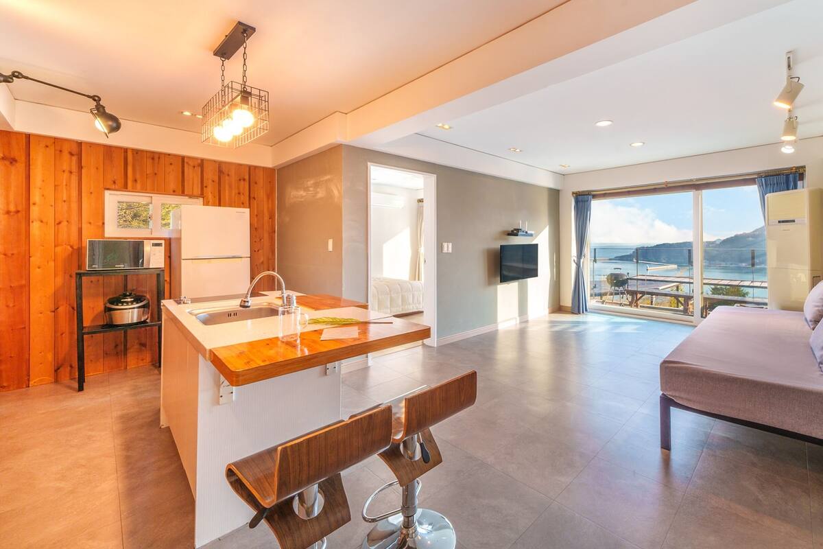 A modern kitchen with wooden accents, a dining area with a bench, and a bedroom with a view.