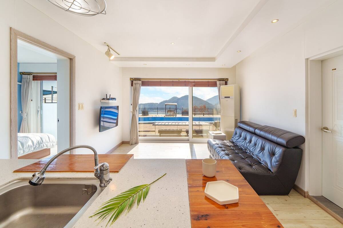 A modern hotel room with a balcony, a sofa, a dining table, and a kitchen area.