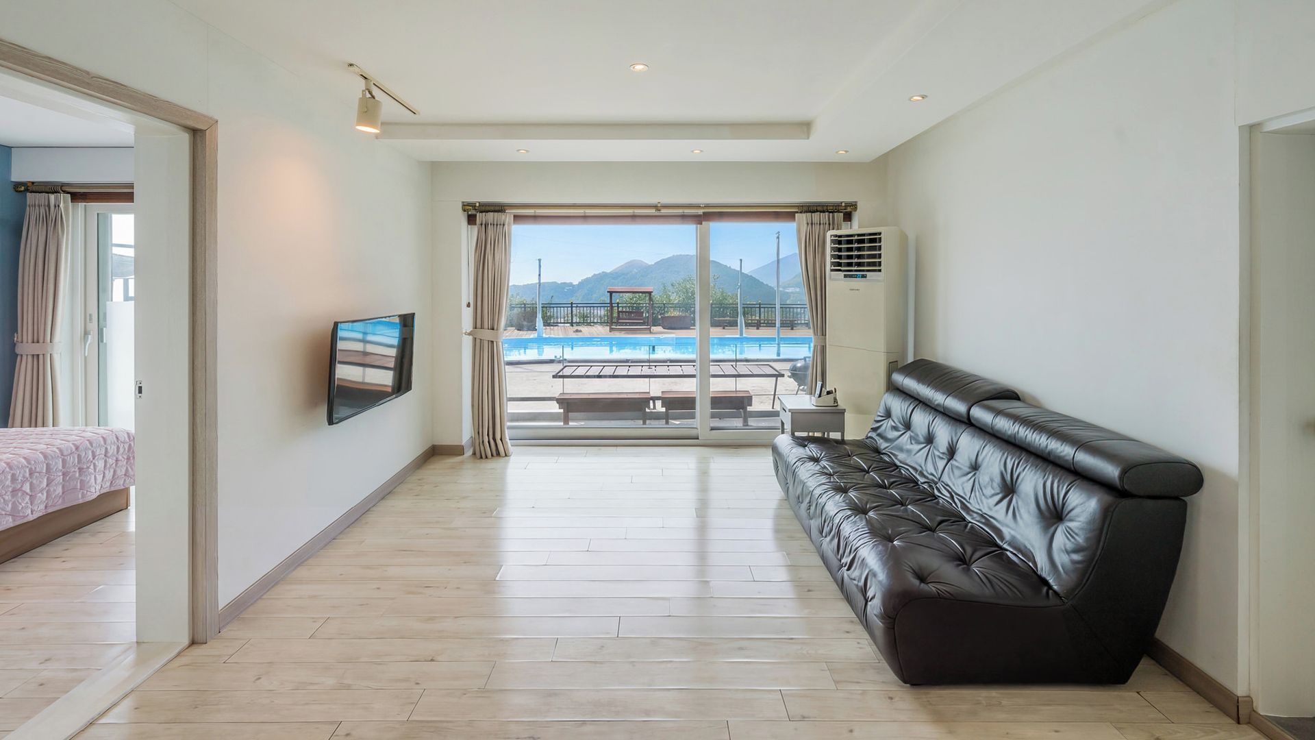 A spacious room with a large window, a flat-screen TV, a leather sofa, and a pool view.