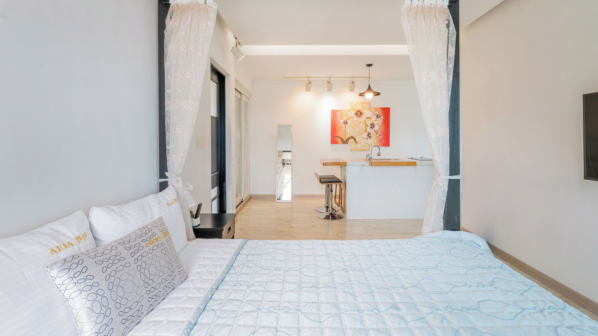 A bedroom with a bed, a nightstand, a window with curtains, a bathroom with a sink and mirror, and a hallway leading to another room.
