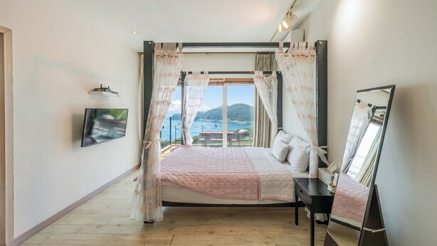 A bedroom with a bed, a TV, a bedside table, and a large window with a view of the sea. - Namhae Aria Spa Pension (Namhae)