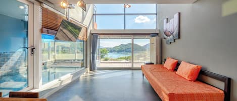 A modern hotel room with a large sliding glass door leading to a balcony, a sofa with patterned cushions, and a view of a mountainous landscape.