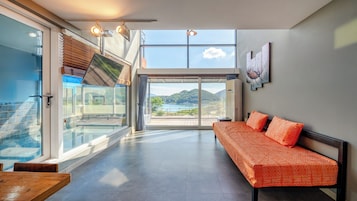A modern hotel room with a large sliding glass door leading to a balcony, a sofa with patterned cushions, and a view of a mountainous landscape.