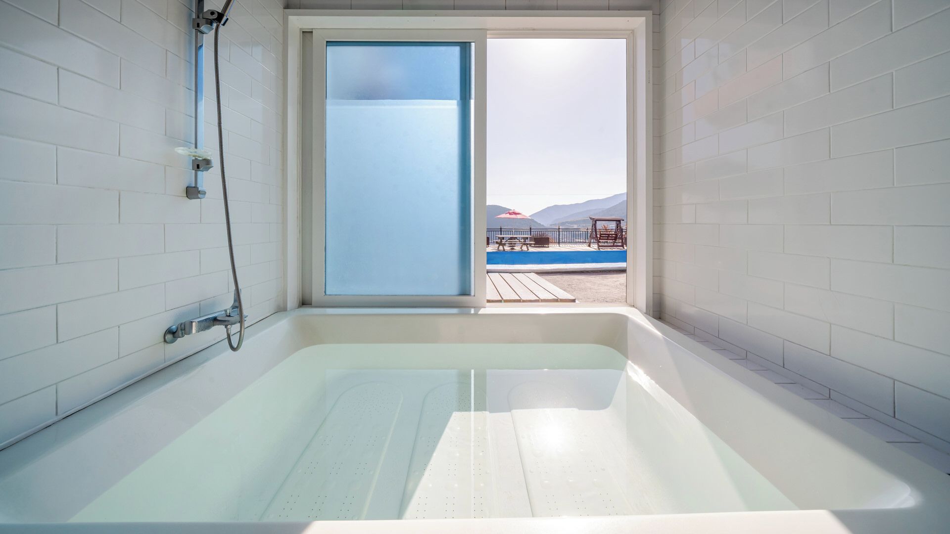 A modern bathroom with a bathtub, a shower, and a large window offering a view of a pool and mountains.