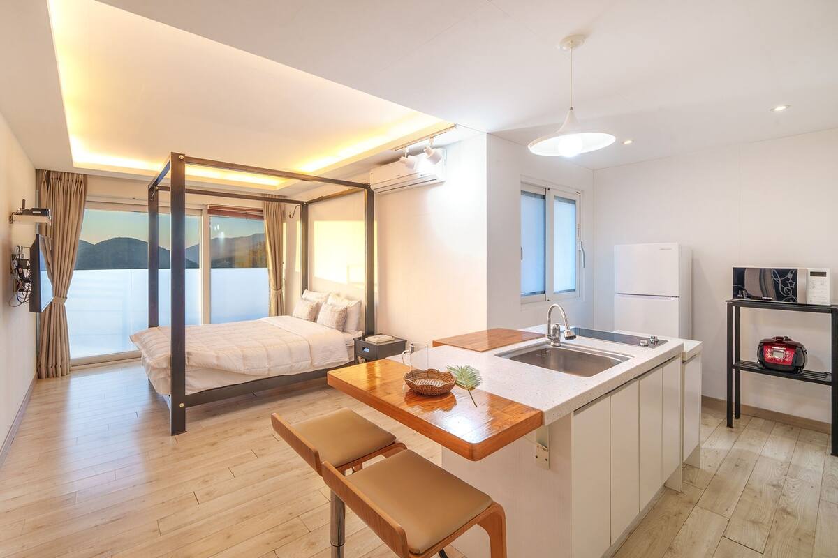 A modern hotel room with a balcony, a bed, a kitchenette, and a dining area.