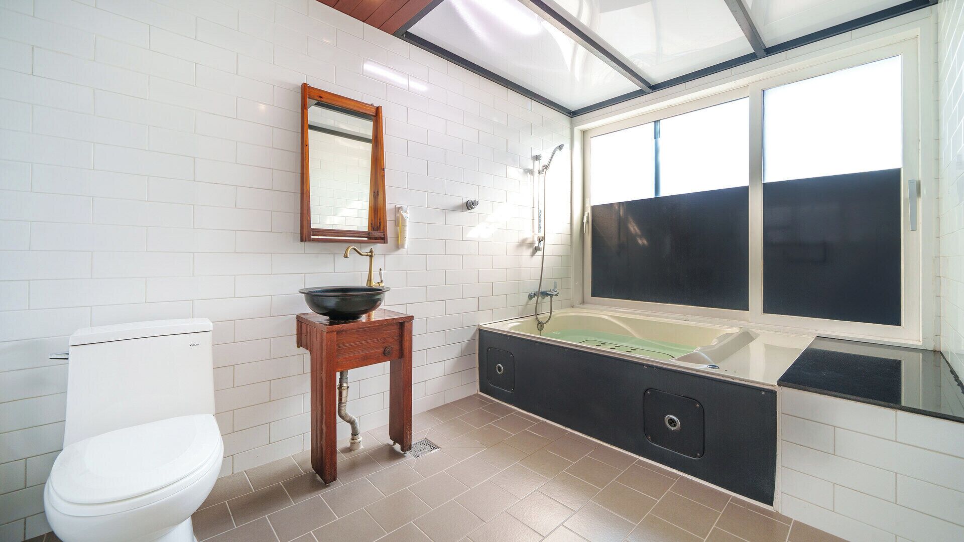 A bathroom with a white toilet, a wooden vanity with a black basin, a large mirror, a bathtub, and a window.