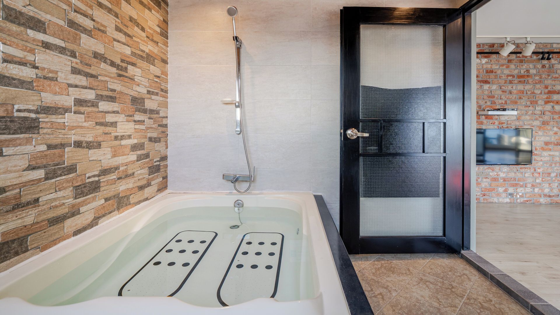 A modern bathroom with a stone wall, a bathtub with foot jets, a showerhead, and a door with a screen.