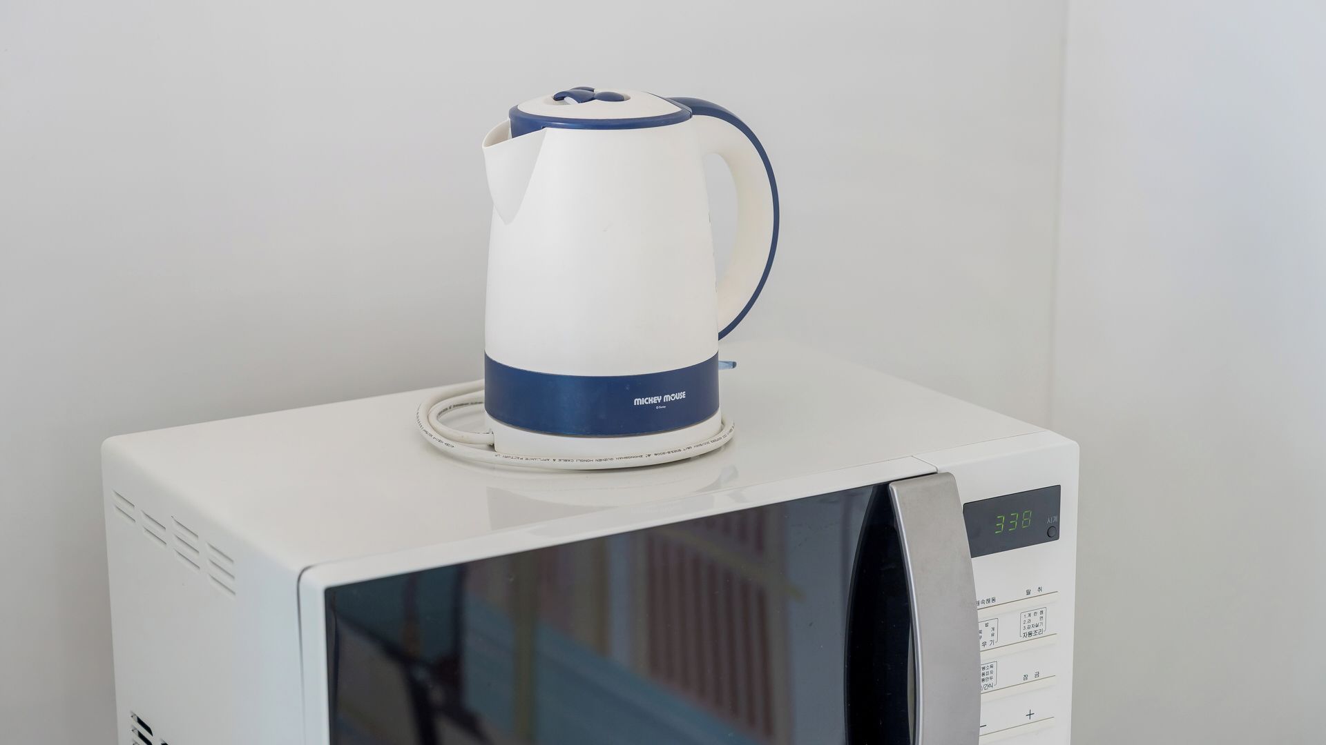 A white microwave with a digital display and a white kettle with a blue base on top.