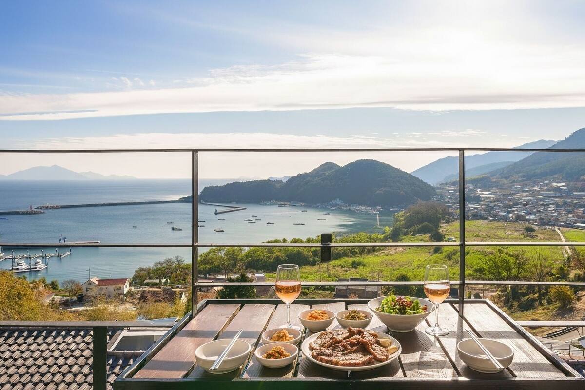 A table with food and drinks, overlooking a coastal view with boats and a distant town.