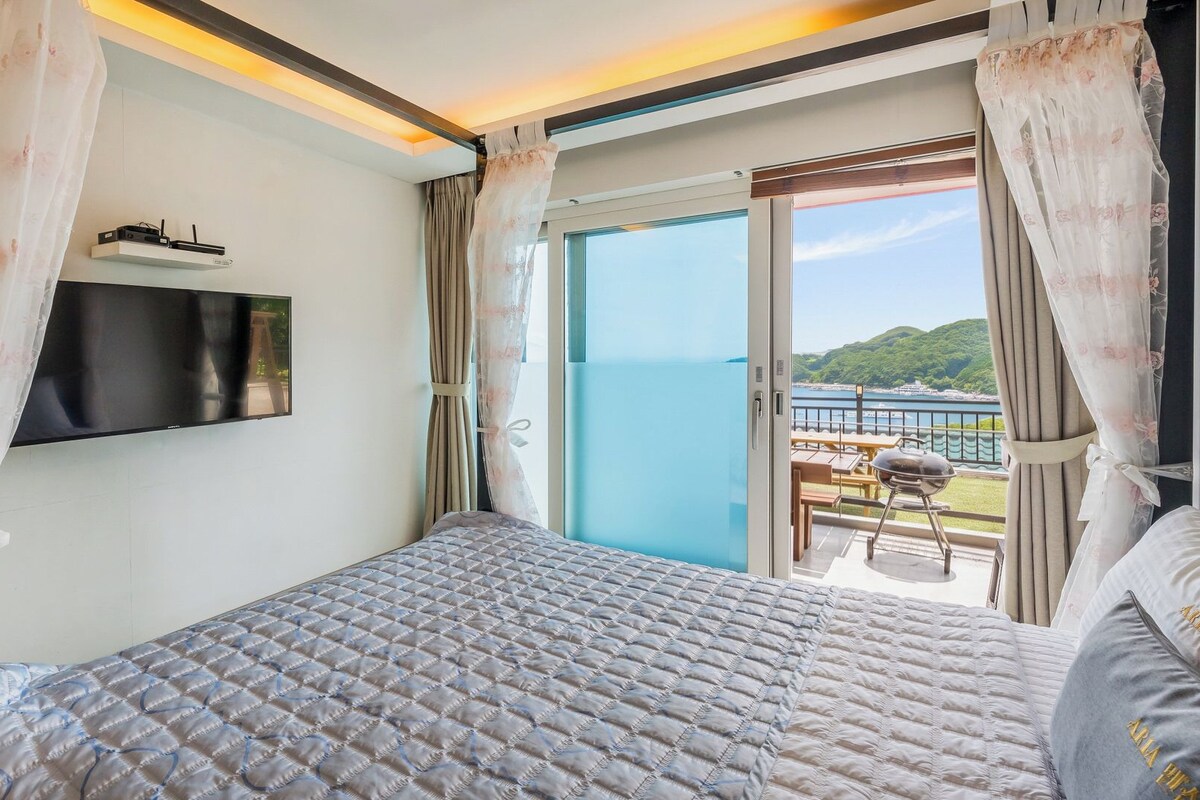 Habitación de hotel con cama, televisión de pantalla plana, balcón con vista al mar y mesa con parrilla.