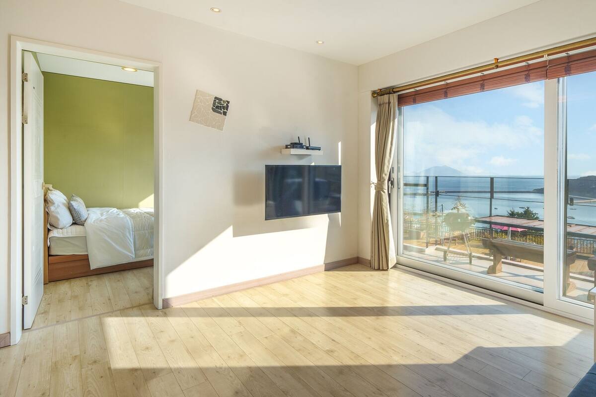 A modern hotel room with a large balcony, a bed, a TV, and a view of the sea.