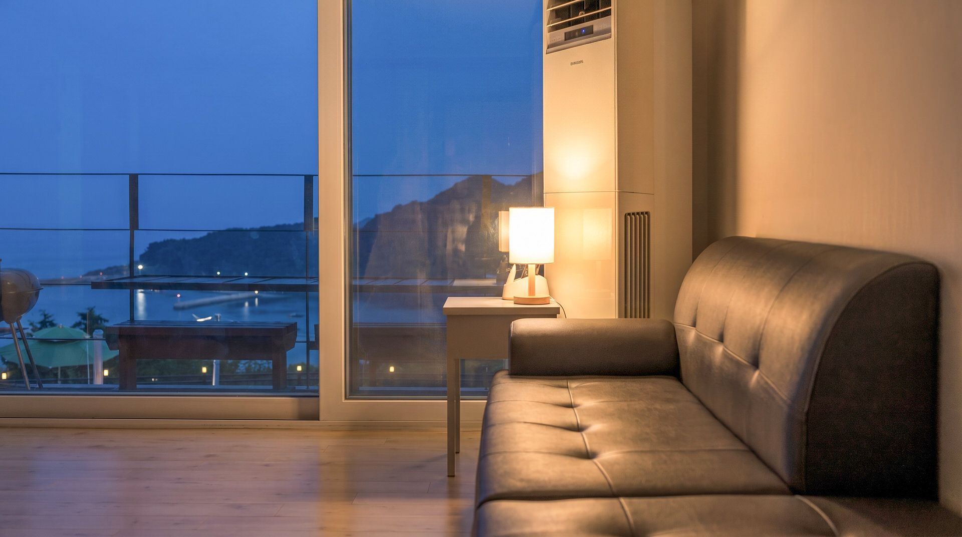 Un salón moderno con un sofá de cuero, una mesita con una lámpara y vistas a una masa de agua a través de amplios ventanales.