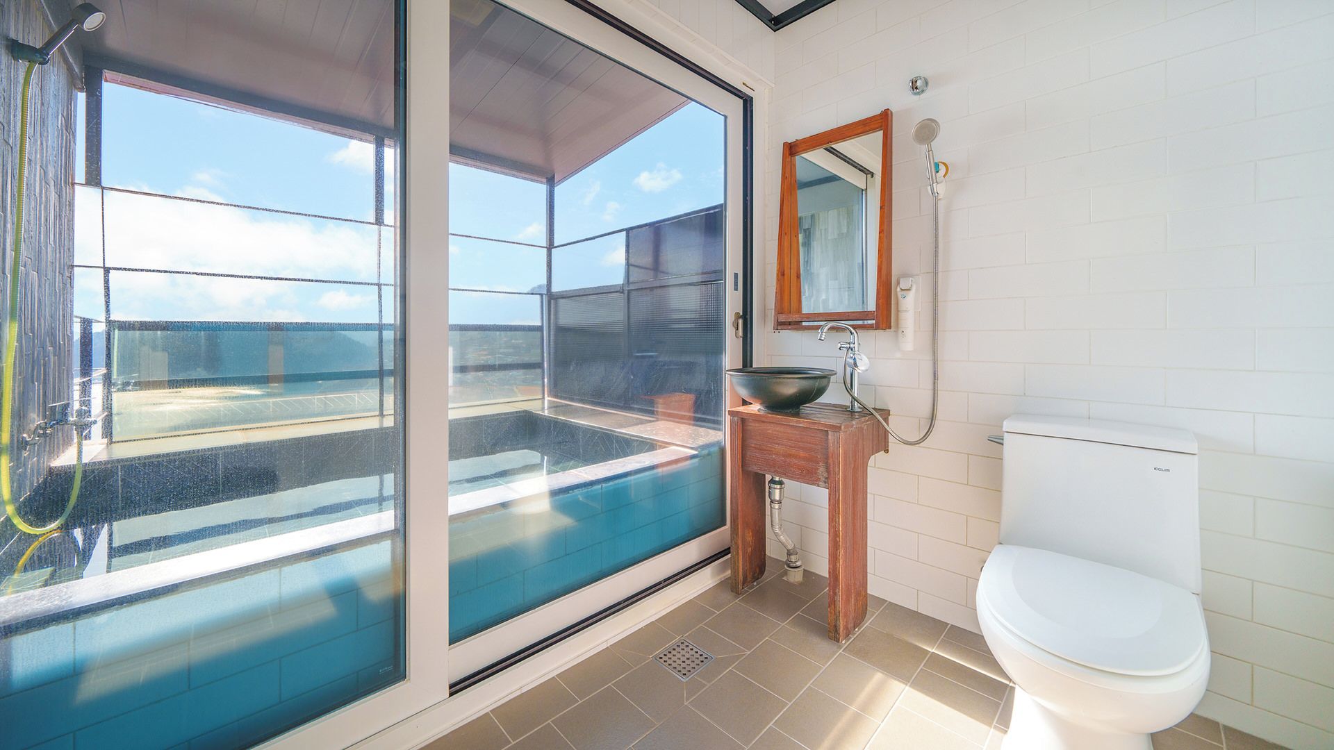 A modern bathroom with a large glass door leading to a balcony, a wooden vanity, a white toilet, and a shower.