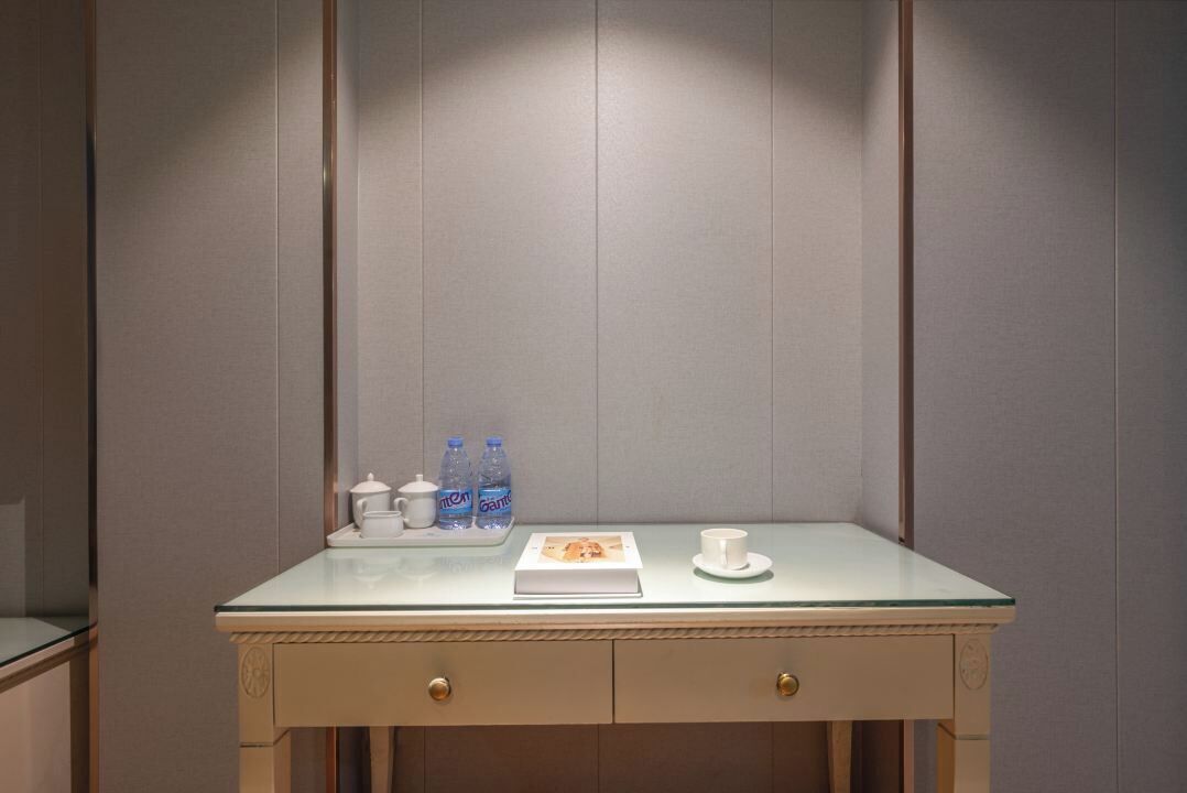 Una habitación pequeña y bien iluminada con una mesa de vidrio, dos cajones y una bandeja con botellas de agua, una tetera y una taza.